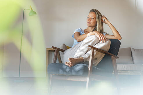 Nachdenkliche Frau mit Hand im Haar, die wegschaut, während sie auf einem Stuhl zu Hause sitzt - SBOF03445