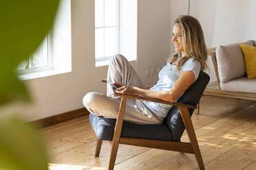 Lächelnde blonde Frau, die wegschaut, während sie mit einem digitalen Tablet auf einem Sessel zu Hause sitzt - SBOF03449