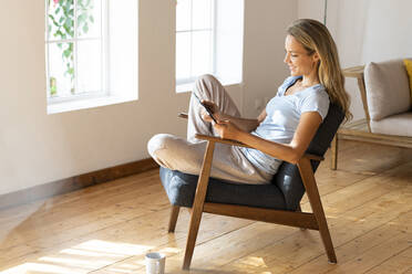 Smiling woman using digital tablet while sitting on chair in living room - SBOF03450