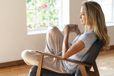 Frau träumt, während sie eine Kaffeetasse auf einem Stuhl zu Hause hält - SBOF03454