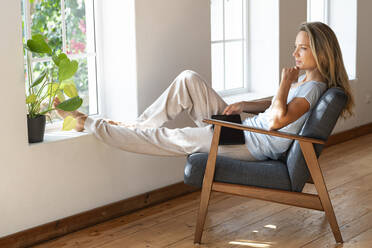 Mid adult woman with hand on chin day dreaming while sitting at window in living room - SBOF03457