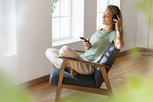Frau hört Musik über Kopfhörer und hält ihr Handy auf einem Sessel im Wohnzimmer - SBOF03468