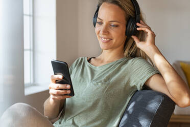 Smiling woman listening music through headphones while using mobile phone at home - SBOF03470