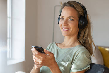 Smiling woman with headphones holding smart phone at home - SBOF03471