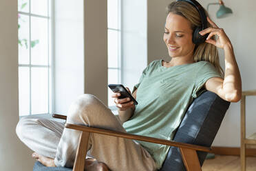 Lächelnde Frau mit Kopfhörern, die ein Smartphone benutzt, während sie auf einem Stuhl im Wohnzimmer sitzt - SBOF03474