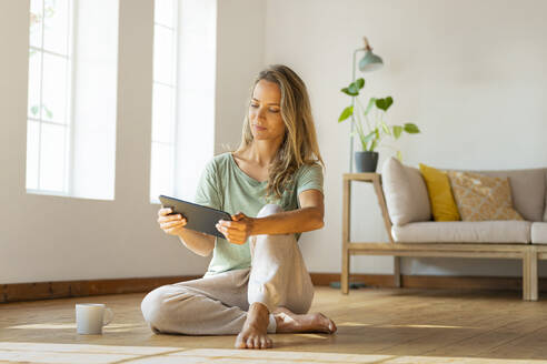 Frau in Loungewear schaut auf ein digitales Tablet, während sie im Wohnzimmer sitzt - SBOF03478