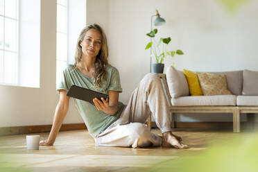 Woman in loungewear holding digital tablet while sitting on hardwood floor at home - SBOF03481