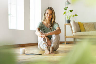 Selbstbewusste Frau in Loungewear sitzt zu Hause mit digitalem Tablet und Kaffeetasse auf dem Boden - SBOF03482