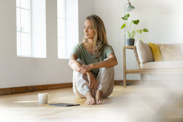 Blond woman day dreaming while sitting by digital tablet and coffee mug on floor in living room - SBOF03483