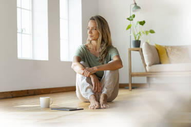 Blonde Frau träumt, während sie mit digitalem Tablet und Kaffeetasse auf dem Boden im Wohnzimmer sitzt - SBOF03483