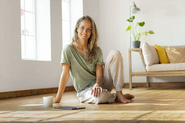 Happy woman sitting on floor by digital tablet and mug at home - SBOF03484