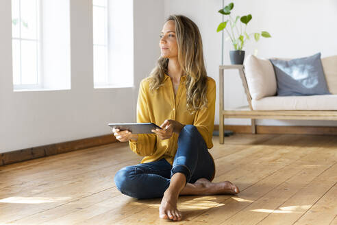 Frau mit digitalem Tablet, die wegschaut, während sie auf dem Boden im Wohnzimmer sitzt - SBOF03490