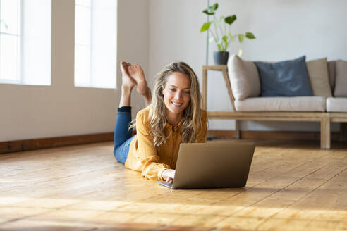 Smiling woman using laptop while lying on harwood floor in living room - SBOF03492
