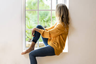 Woman sitting on windowsill while looking through window in living room - SBOF03496