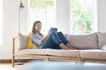 Lächelnde Frau mit Brille, die ein digitales Tablet auf einer Couch im Wohnzimmer hält - SBOF03497
