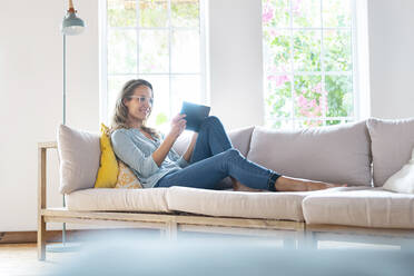 Lächelnde Frau schaut auf ein digitales Tablet, während sie sich auf dem Sofa entspannt - SBOF03498