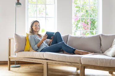 Lächelnde schöne Frau, die wegschaut und ein digitales Tablet auf dem Sofa im Wohnzimmer hält - SBOF03499