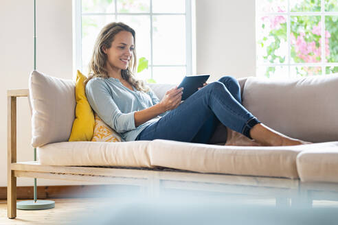 Lächelnde Frau in Freizeitkleidung schaut auf ein digitales Tablet, während sie auf der Couch sitzt - SBOF03501