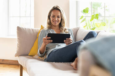 Contented woman looking at digital tablet while sitting on sofa in living room - SBOF03502