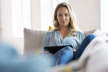 Blond woman with digital tablet sitting on couch at home - SBOF03503