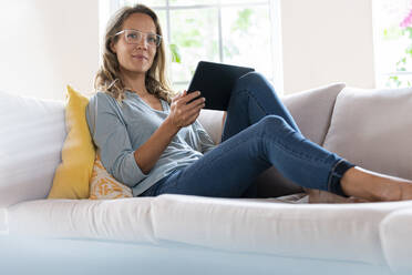 Beautiful woman with digital tablet relaxing on sofa in living room - SBOF03504