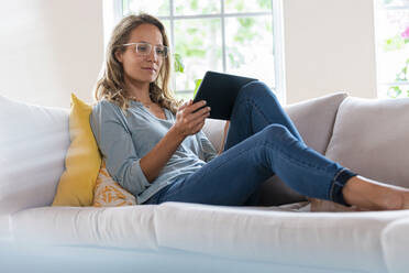 Woman with eyeglasses using digital tablet while sitting on sofa at home - SBOF03505