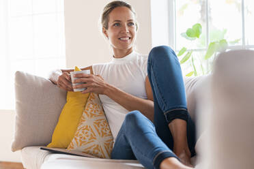 Smiling woman day dreaming while having coffee on sofa at home - SBOF03511