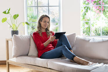 Blonde Frau mit digitalem Tablet sitzt auf dem Sofa im Wohnzimmer - SBOF03514