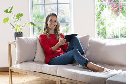 Zufriedene Frau mit digitalem Tablet, die zu Hause auf der Couch sitzt - SBOF03515
