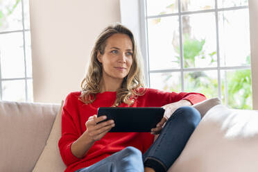 Smiling woman relaxing while holding digital tablet on couch in living room - SBOF03517