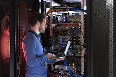 Male IT expertise with laptop looking away while standing at rack in data center - FKF04117