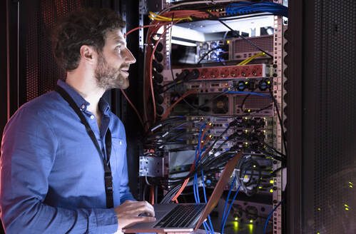 Männlicher IT-Fachmann schaut weg, während er einen Laptop an einem Server-Rack im Rechenzentrum hält - FKF04120
