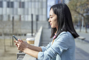 Businesswoman text messaging through smart phone while leaning on railing - VEGF04123