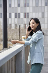 Lächelnde Geschäftsfrau, die mit ihrem Handy telefoniert und sich auf ein Geländer stützt - VEGF04126