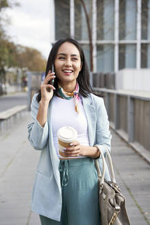 Smiling businesswoman talking on mobile phone while walking on footpath - VEGF04129