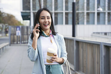 Cheerful businesswoman with coffee mug talking on mobile phone while walking on footpath - VEGF04130