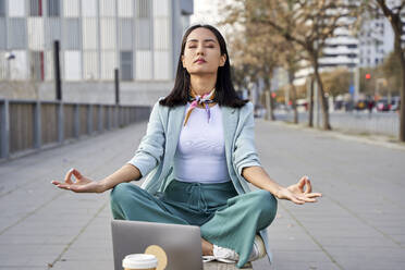 Young businesswoman with laptop meditating on bench at footpath - VEGF04136