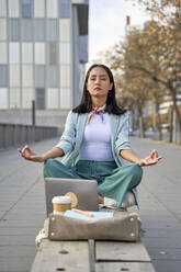 Businesswoman with laptop meditating while sitting on bench at footpath - VEGF04137