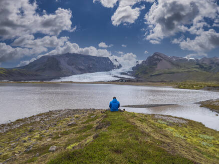 Mittlerer erwachsener Mann, der vor einer Lagune sitzend die Aussicht betrachtet, Breidamerkurjokull-Gletscher - LAF02703