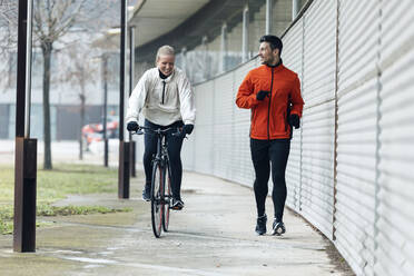 Lächelnde junge Frau, die mit dem Fahrrad an einem männlichen Freund vorbeifährt, der auf dem Fußweg joggt - JSRF01460