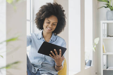 Lächelnde Frau, die ein digitales Tablet benutzt, während sie am Fenster sitzt - SBOF03520