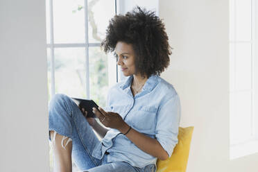 Nachdenkliche Frau, die ein digitales Tablet hält, während sie auf der Fensterbank sitzt - SBOF03522