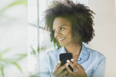 Lächelnde Frau mit Mobiltelefon, die durch das Fenster ihres Hauses schaut - SBOF03527