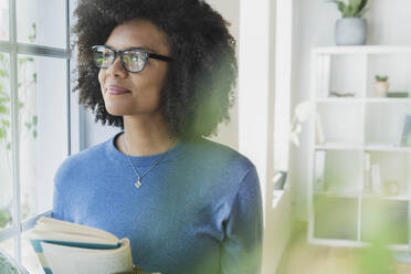 Woman with eyeglasses smiling while looking through window at home - SBOF03530