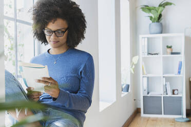 Junge Frau liest ein Buch, während sie zu Hause am Fenster sitzt - SBOF03531