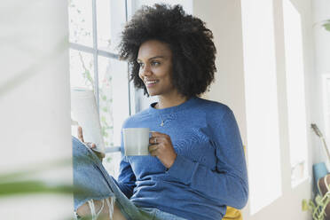 Frau mit Kaffeetasse und Buch am Fenster zu Hause - SBOF03534