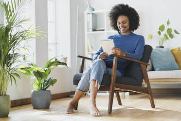 Smiling woman reading book while sitting on armchair at home - SBOF03537