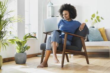 Smiling woman using laptop while sitting on armchair at home - SBOF03541