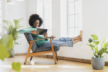 Lächelnde Frau, die ein digitales Tablet benutzt, während sie zu Hause auf einem Sessel sitzt - SBOF03544
