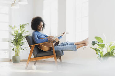 Smiling young woman playing guitar while sitting on armchair at home - SBOF03546